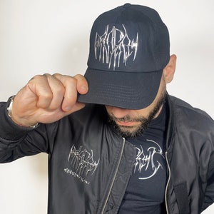 K&ouml;nnte beinhalten: Eine schwarze Baseballkappe mit einem silbernen Metallic-Logo, das in gotischer Schrift "DEADLY" lautet. Die Kappe wird von einer Person getragen, die eine schwarze Bomberjacke mit einem &auml;hnlichen Logo auf der linken Seite der Jacke tr&auml;gt.