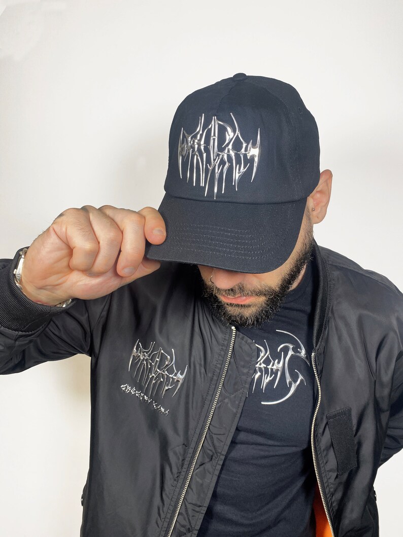 K&ouml;nnte beinhalten: Eine schwarze Baseballkappe mit einem silbernen Metallic-Logo, das in gotischer Schrift "DEADLY" lautet. Die Kappe wird von einer Person getragen, die eine schwarze Bomberjacke mit einem &auml;hnlichen Logo auf der linken Seite der Jacke tr&auml;gt.