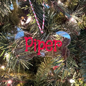 May include: A clear plastic dog bone ornament with the name "Piper" written in red on it. The ornament is hanging on a Christmas tree with red and black plaid ornaments.