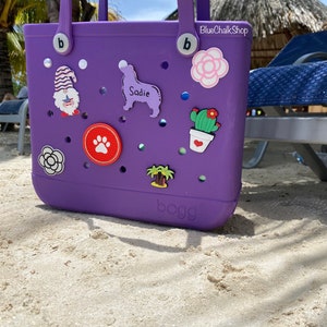 May include: Purple Bogg bag with various colorful charms, including a flower, a cactus, a palm tree, a paw print, a gnome, and a dog silhouette with the name "Sadie". The bag has two handles and the text "BlueChalkShop" and "bogg" are visible.
