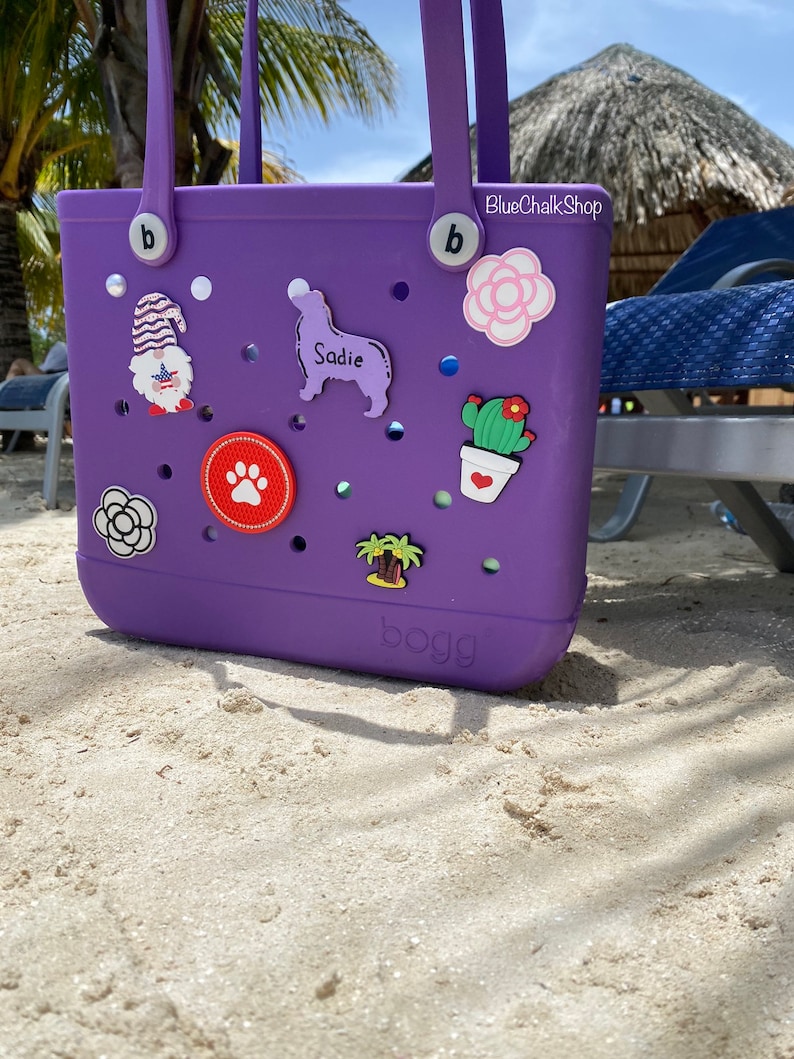 May include: Purple Bogg bag with various colorful charms, including a flower, a cactus, a palm tree, a paw print, a gnome, and a dog silhouette with the name "Sadie". The bag has two handles and the text "BlueChalkShop" and "bogg" are visible.
