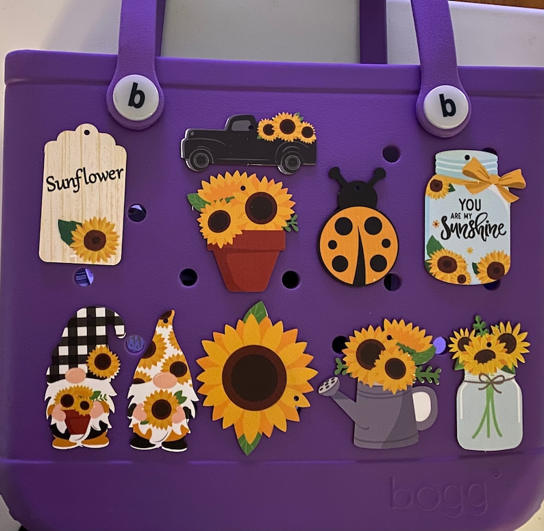 May include: Purple tote bag with various colorful stickers, including a sunflower, a truck, a ladybug, a watering can, a gnome, and a jar with the text "You are my sunshine".