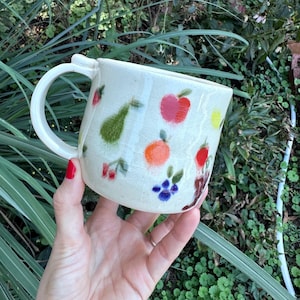 May include: A cream-colored ceramic mug with a glossy, crackled glaze finish. The exterior is decorated with small, hand-painted illustrations of various fruits including a green pear, a red apple, an orange, cherries, and blue berries.