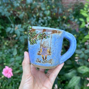 May include: A light blue ceramic mug with a speckled finish featuring a hand-painted illustration of a person sitting on a swing hanging from a tree branch. The text "Be Still" is written in brown script below the swing illustration.