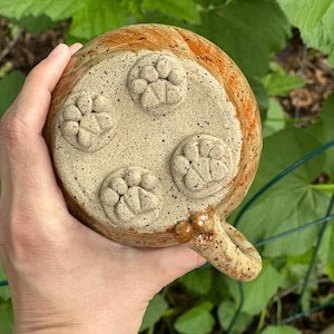 May include: A handmade ceramic mug with a speckled beige base and a paw print design. The rim is a warm orange-brown colour. The mug has a rounded handle and is held in a hand against a background of green foliage.