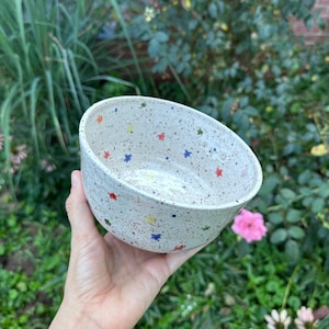 May include: A round ceramic bowl with a speckled off-white base featuring small, colorful star-shaped accents in red, blue, yellow, and green scattered across the interior and exterior surfaces of the vessel, showcasing a smooth, glazed finish.