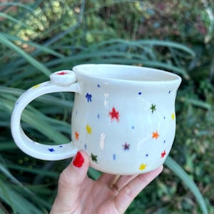 May include: A white ceramic mug with a rounded body and a curved handle, featuring a hand-painted pattern of small, multi-colored stars and dots in red, blue, yellow, green, and orange scattered across the exterior surface.