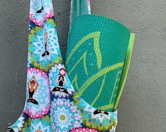 May include: A white and blue yoga bag with a green yoga mat inside. The bag has a pattern of colorful mandalas with images of people in yoga poses.
