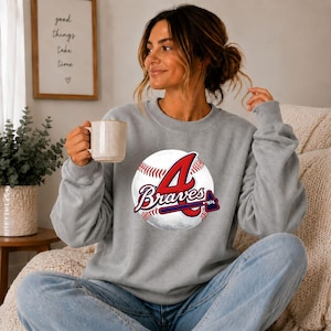 May include: A heather grey crewneck sweatshirt featuring a graphic of a white baseball with red stitching and the red and navy blue text "A Braves" with a tomahawk icon. The sweatshirt has long sleeves and a ribbed collar, cuffs, and hem.