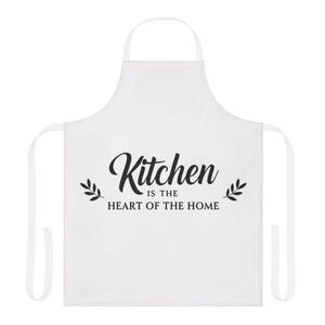 以下が含まれることがあります： 白いキッチンエプロン。黒い筆記体で「Kitchen is the Heart of the Home」と書かれています。エプロンにはビブ、ウエストタイ、ネックループが付いています。テキストの両側に葉の小枝があります。