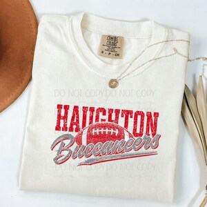 May include: Cream-colored t-shirt with the text "HAUGHTON Buccaneers" in red and gray, featuring a football graphic. The shirt has a Comfort Colors tag and a small necklace. The shirt is folded.