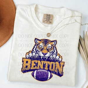 May include: Cream-colored t-shirt featuring a tiger graphic with the word "BENTON" in orange and purple. The tiger is depicted with sharp teeth and claws, and a football is below the text. The shirt has a crew neck and short sleeves.