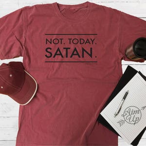 May include: A maroon t-shirt with the text "NOT. TODAY. SATAN." printed in black. The shirt is laid flat on a white wooden surface with a black and white notebook, a red baseball cap, and a black travel mug.