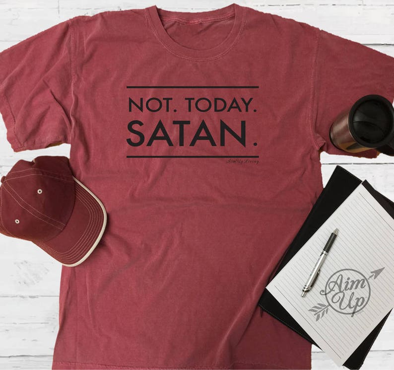 May include: A maroon t-shirt with the text "NOT. TODAY. SATAN." printed in black. The shirt is laid flat on a white wooden surface with a black and white notebook, a red baseball cap, and a black travel mug.