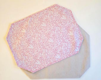 May include: Two octagonal placemats. The top placemat features a white fabric with a dense, repeating pattern of thin red heart outlines. The bottom placemat is made of a plain, solid light beige or oatmeal-colored fabric.