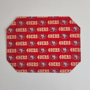 May include: Red and gold placemat featuring the San Francisco 49ers logo and team name repeated across the surface. The placemat has an octagonal shape with rounded corners. The background is red.