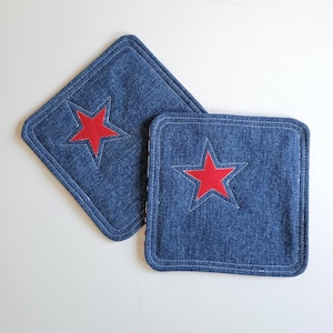 May include: Two denim square coasters with a red star applique. The coasters have white stitching around the edges. The denim is a medium blue color.