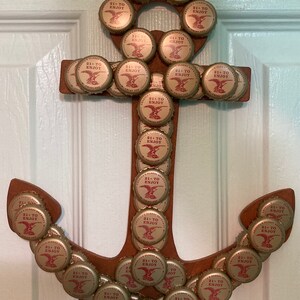 May include: A wooden anchor decorated with numerous beer bottle caps. The caps are gold and red, with the text "21+ TO ENJOY" and an eagle emblem. The anchor is hanging on a white door.
