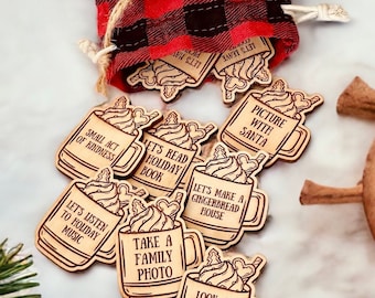 Puede incluir: Un conjunto de 10 adornos navideños de madera con forma de taza con actividades festivas de Navidad escritas en ellos. Los adornos están en una bolsa de cordón de cuadros rojos y negros.