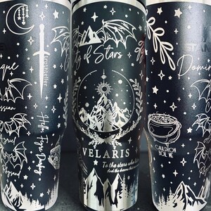 May include: A matte black stainless steel tumbler featuring white laser-engraved illustrations of mountains, wings, stars, a sword, and a cauldron. Text includes "City of Stars," "VELARIS," "To the stars who listen," "Dominique," "Cassian," "Azriel," "Rhysand," and "Cauldron Boil Me."