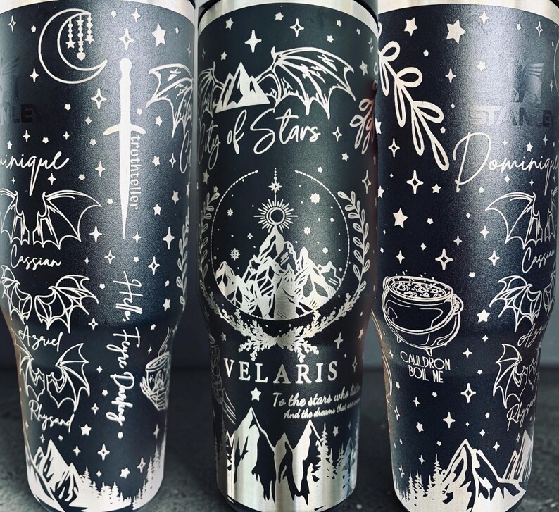 May include: A matte black stainless steel tumbler featuring white laser-engraved illustrations of mountains, wings, stars, a sword, and a cauldron. Text includes "City of Stars," "VELARIS," "To the stars who listen," "Dominique," "Cassian," "Azriel," "Rhysand," and "Cauldron Boil Me."
