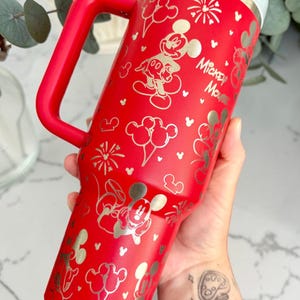 May include: A red insulated tumbler with a handle, featuring gold-colored Mickey Mouse illustrations and the text "Mickey Mouse." The design includes Mickey Mouse, balloons, and fireworks. The tumbler is designed to keep beverages cold or hot.
