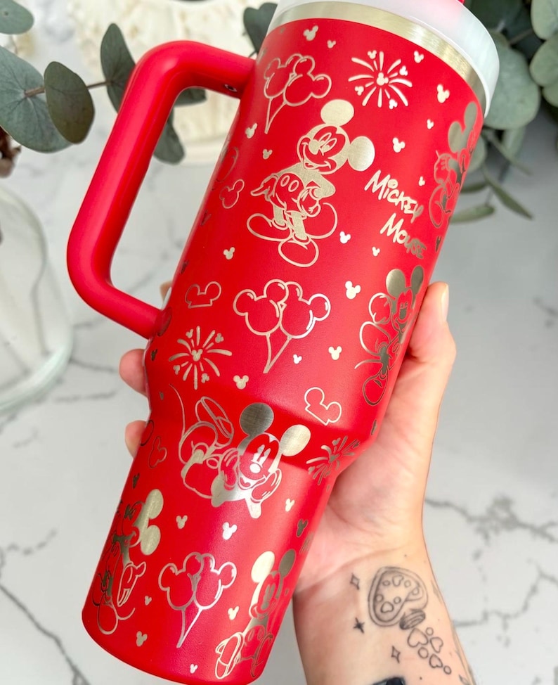 May include: A red insulated tumbler with a handle, featuring gold-colored Mickey Mouse illustrations and the text "Mickey Mouse." The design includes Mickey Mouse, balloons, and fireworks. The tumbler is designed to keep beverages cold or hot.