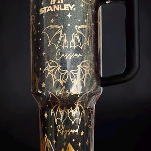 May include: A black glittery Stanley tumbler with a gold-toned design featuring bat wings, stars, a crescent moon, and mountain silhouettes. The names Cassian, Azriel, and Rhysand are printed in gold script between the bat wing illustrations.