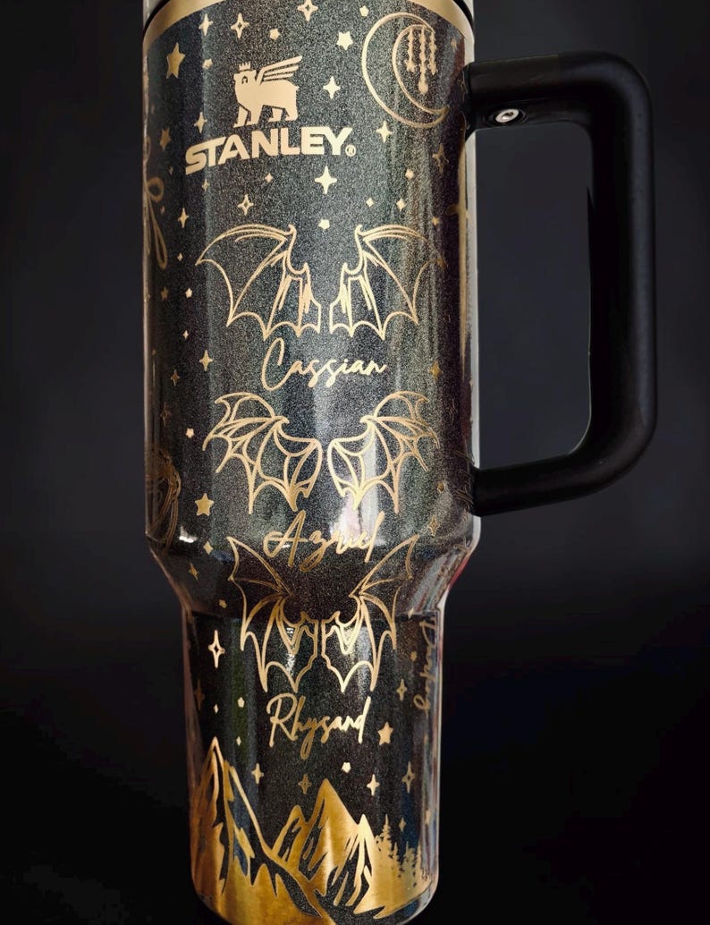 May include: A black glittery Stanley tumbler with a gold-toned design featuring bat wings, stars, a crescent moon, and mountain silhouettes. The names Cassian, Azriel, and Rhysand are printed in gold script between the bat wing illustrations.