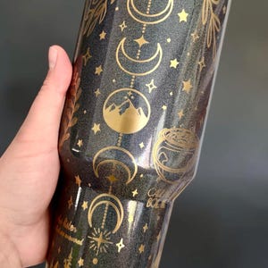 May include: A dark gray glitter tumbler featuring gold metallic designs of moon phases, stars, mountain peaks, and a cauldron. The text "CALL BACK" and "do listen" is printed in gold. The base of the tumbler features a gold forest silhouette.