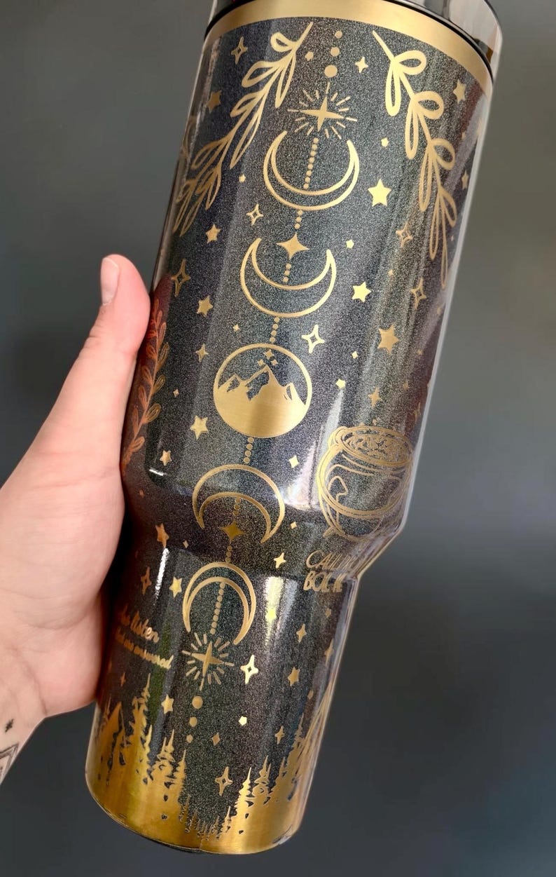 May include: A dark gray glitter tumbler featuring gold metallic designs of moon phases, stars, mountain peaks, and a cauldron. The text "CALL BACK" and "do listen" is printed in gold. The base of the tumbler features a gold forest silhouette.