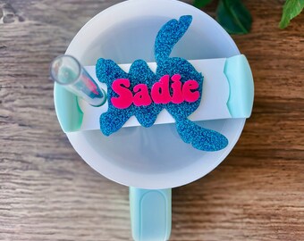Könnte beinhalten: Eine türkisblaue und weiße Reise-Trinkflasche mit Deckel und Strohhalm. Der Deckel hat einen blauen Glitzer-Aufkleber mit einer Comicfigur und dem Namen "Sadie" in Pink.