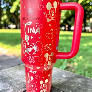 May include: A red Stanley tumbler with a large red handle, featuring silver Mickey Mouse designs and the name "Tina". The tumbler has a stainless steel rim and is decorated with various Mickey Mouse illustrations and fireworks.