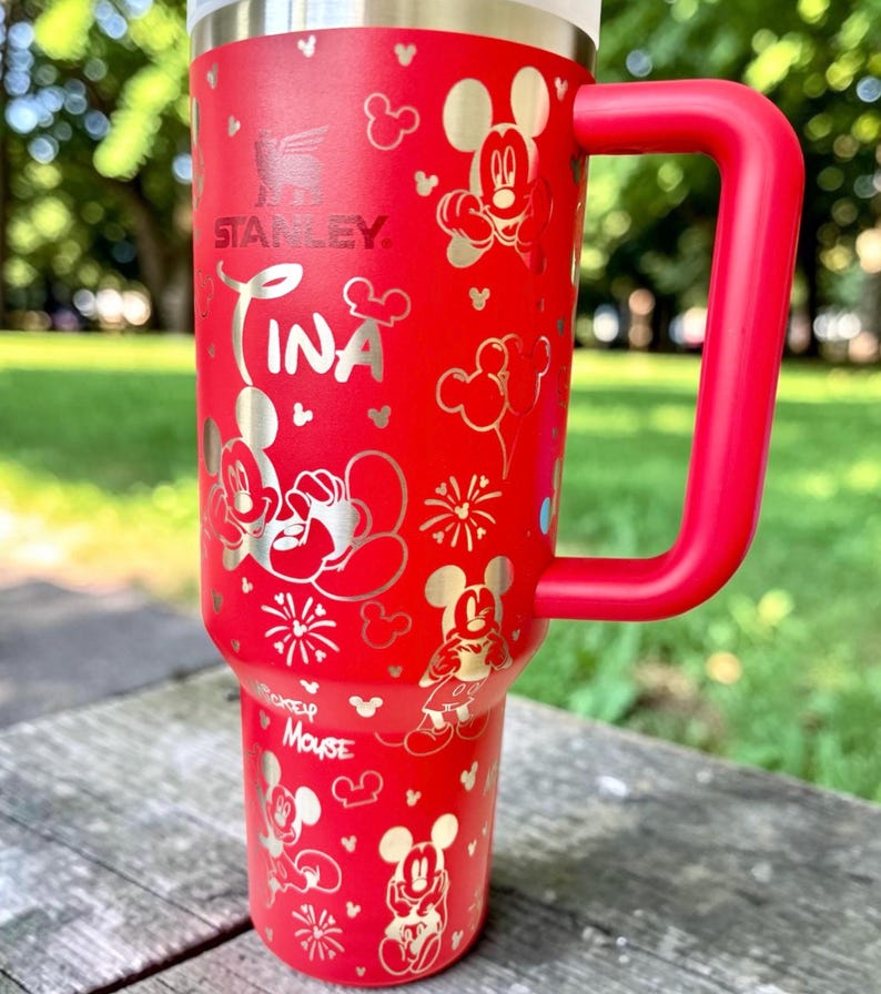 May include: A red Stanley tumbler with a large red handle, featuring silver Mickey Mouse designs and the name "Tina". The tumbler has a stainless steel rim and is decorated with various Mickey Mouse illustrations and fireworks.