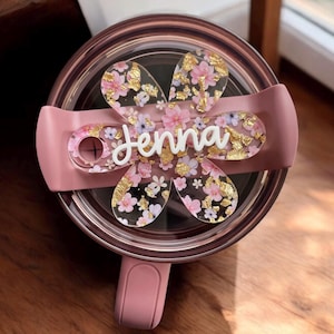 May include: A pink travel mug with a floral design and the name "Jenna" on a clear plastic lid.