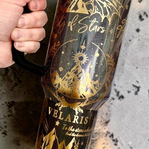 May include: A black glittery tumbler with a handle featuring gold foil designs of mountains, wings, and stars. The text reads City of Stars, VELARIS, and To the stars who listen and the dreams that are answered in a decorative gold script font.