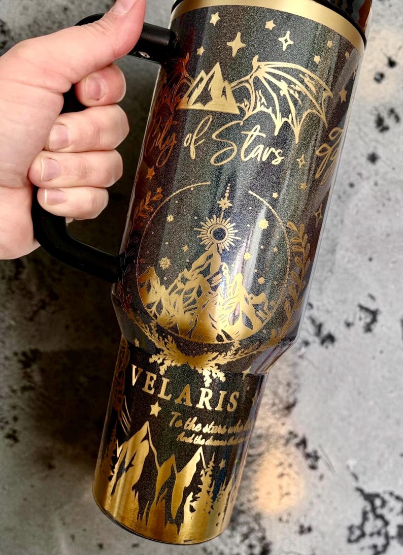 May include: A black glittery tumbler with a handle featuring gold foil designs of mountains, wings, and stars. The text reads City of Stars, VELARIS, and To the stars who listen and the dreams that are answered in a decorative gold script font.