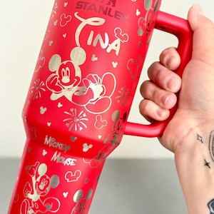 May include: A red Stanley tumbler with a silver Mickey Mouse design and the name "Tina". The tumbler has a red handle and a silver rim. Mickey Mouse and other Disney-themed graphics are printed on the tumbler.