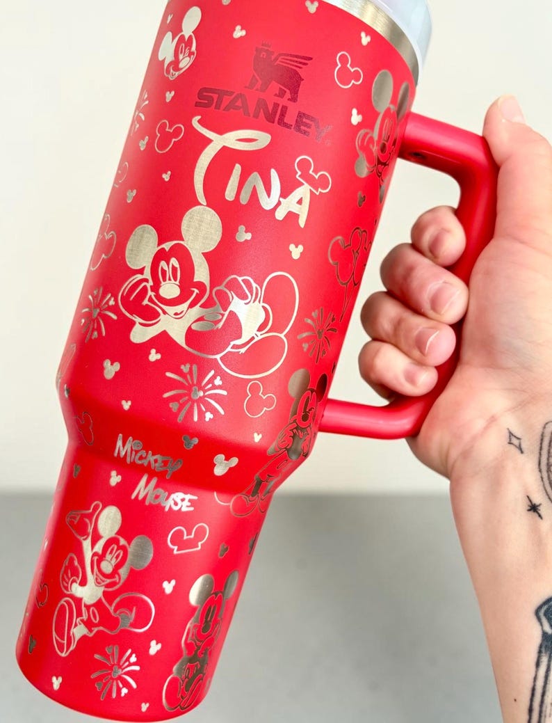 May include: A red Stanley tumbler with a silver Mickey Mouse design and the name "Tina". The tumbler has a red handle and a silver rim. Mickey Mouse and other Disney-themed graphics are printed on the tumbler.