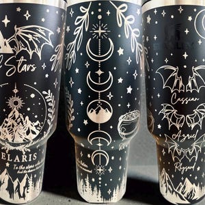 May include: A matte black stainless steel tumbler featuring laser-engraved white illustrations of mountains, bat wings, moon phases, stars, and a cauldron. The text includes City of Stars, Velaris, Cassian, Azriel, and Rhysand in various fonts.