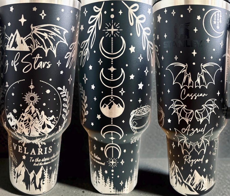 May include: A matte black stainless steel tumbler featuring laser-engraved white illustrations of mountains, bat wings, moon phases, stars, and a cauldron. The text includes City of Stars, Velaris, Cassian, Azriel, and Rhysand in various fonts.