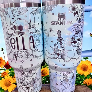 May include: Two light blue Stanley tumblers featuring a Stitch design with floral and tropical leaf accents. One tumbler has the name "Ella" in black script. The other tumbler has the Stanley logo and Stitch playing a guitar. The tumblers have a silver rim and handle.