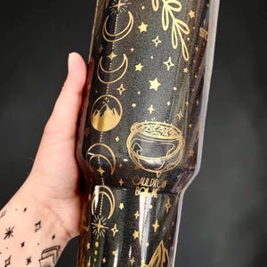 May include: A black glittery tumbler featuring gold metallic graphics of moon phases, stars, a mountain range, and a cauldron. The text CAULDRON BORN is printed in gold. The base of the tumbler features a gold mountain and forest landscape design.