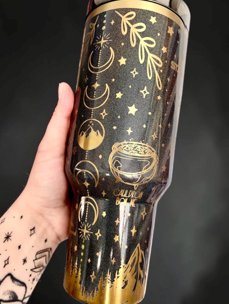 May include: A black glittery tumbler featuring gold metallic graphics of moon phases, stars, a mountain range, and a cauldron. The text CAULDRON BORN is printed in gold. The base of the tumbler features a gold mountain and forest landscape design.
