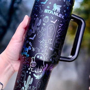 May include: Black Stanley tumbler with a handle, decorated with Halloween-themed illustrations in iridescent colors. The design includes bats, ghosts, cacti, and the Stanley logo.