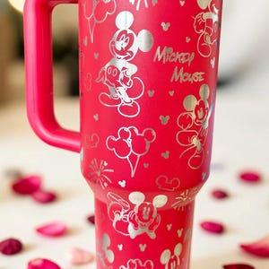 May include: A red insulated tumbler with a handle, featuring a silver Mickey Mouse design. The design includes Mickey Mouse illustrations, balloons, fireworks, and the text "Mickey Mouse".