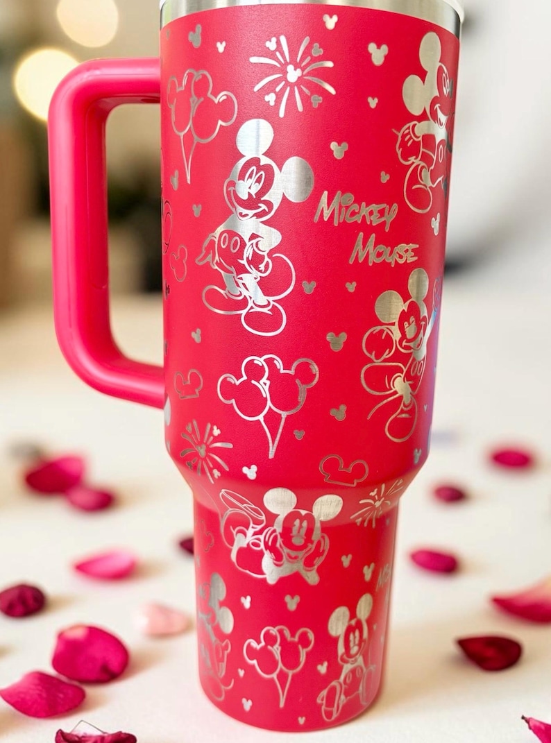 May include: A red insulated tumbler with a handle, featuring a silver Mickey Mouse design. The design includes Mickey Mouse illustrations, balloons, fireworks, and the text "Mickey Mouse".