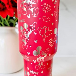 May include: A red insulated tumbler featuring a silver Mickey Mouse design. The tumbler has a variety of Mickey Mouse illustrations, including the character's face, gloves, and poses. The words "Mickey Mouse" are also visible on the tumbler.