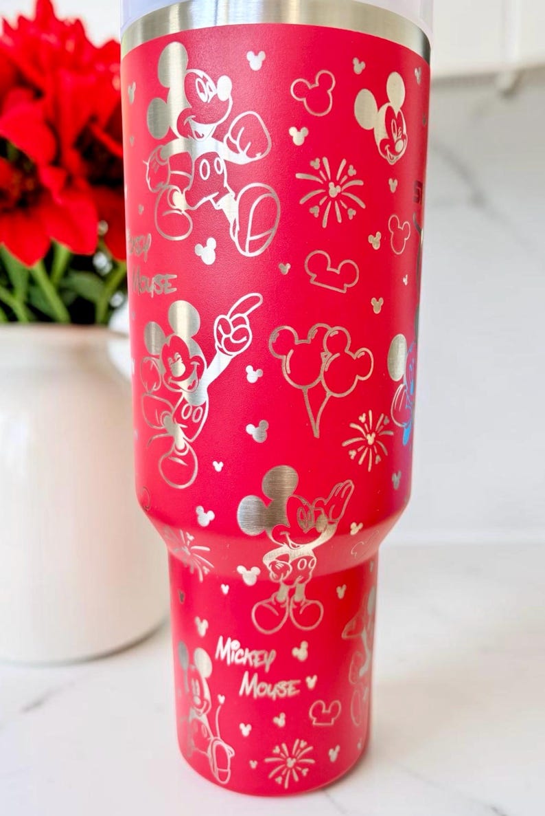 May include: A red insulated tumbler featuring a silver Mickey Mouse design. The tumbler has a variety of Mickey Mouse illustrations, including the character's face, gloves, and poses. The words "Mickey Mouse" are also visible on the tumbler.