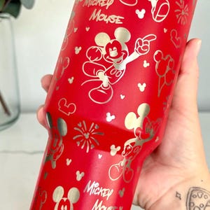 May include: A red tumbler featuring a silver Mickey Mouse design. The tumbler has a silver rim and is covered in Mickey Mouse illustrations, fireworks, and the words "Mickey Mouse".